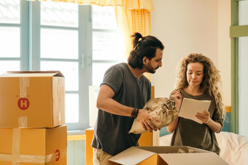 pexels-photo-4247768-4247768 Cheerful young man and woman smiling while unpacking carton boxes with belongings in new apartment during relocation and looking at paper